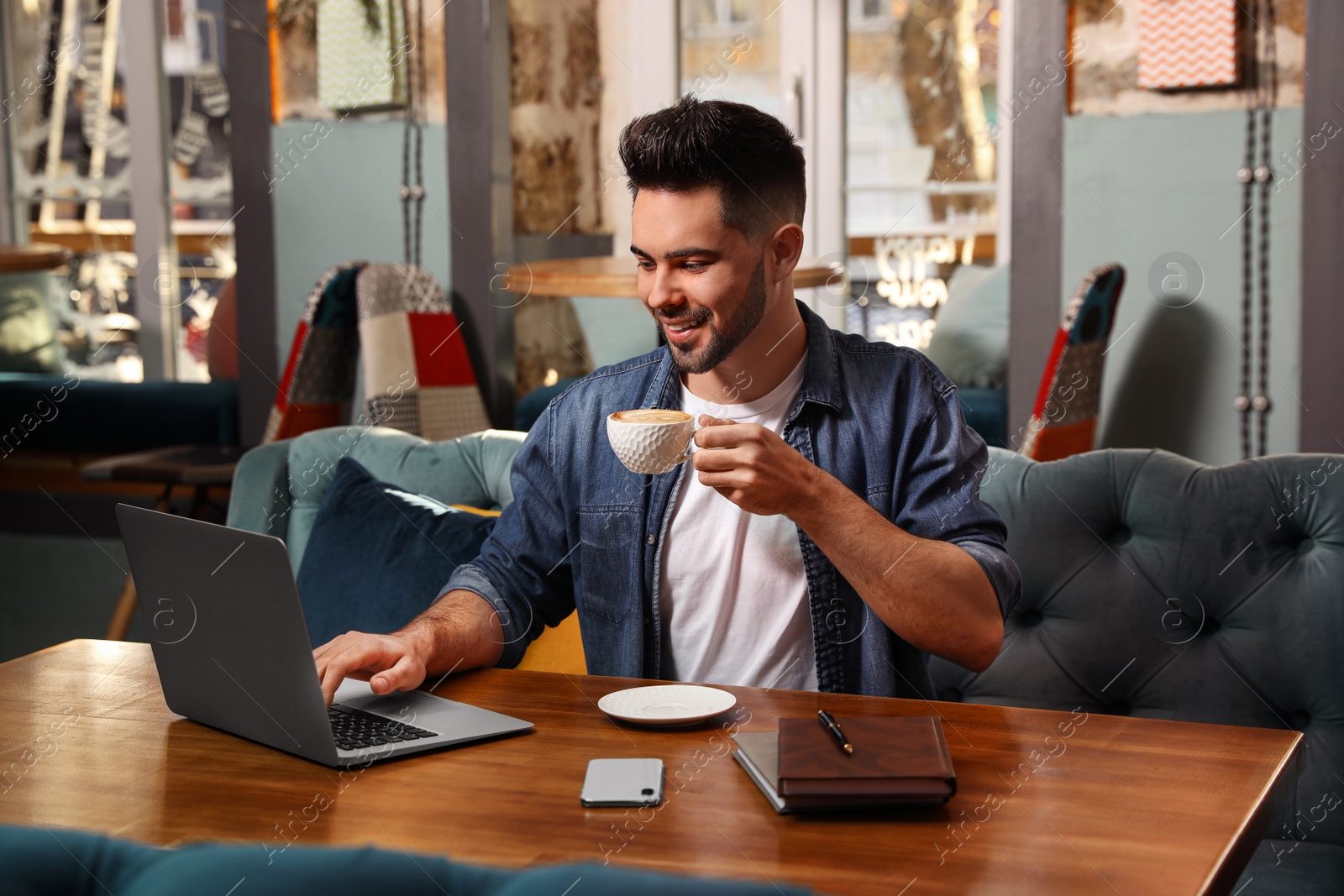 Photo of Young blogger with laptop drinking coffee at table in cafe