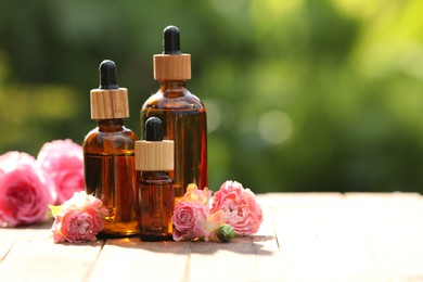 Photo of Bottles of rose essential oil and flowers on wooden table outdoors, space for text