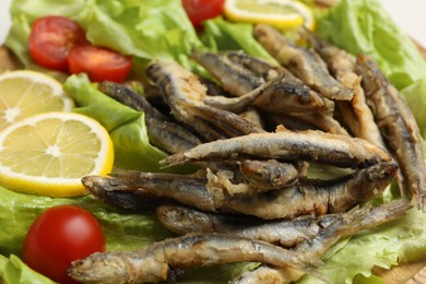 Photo of Delicious fried anchovies with lemon, tomatoes and lettuce leaves as background, closeup