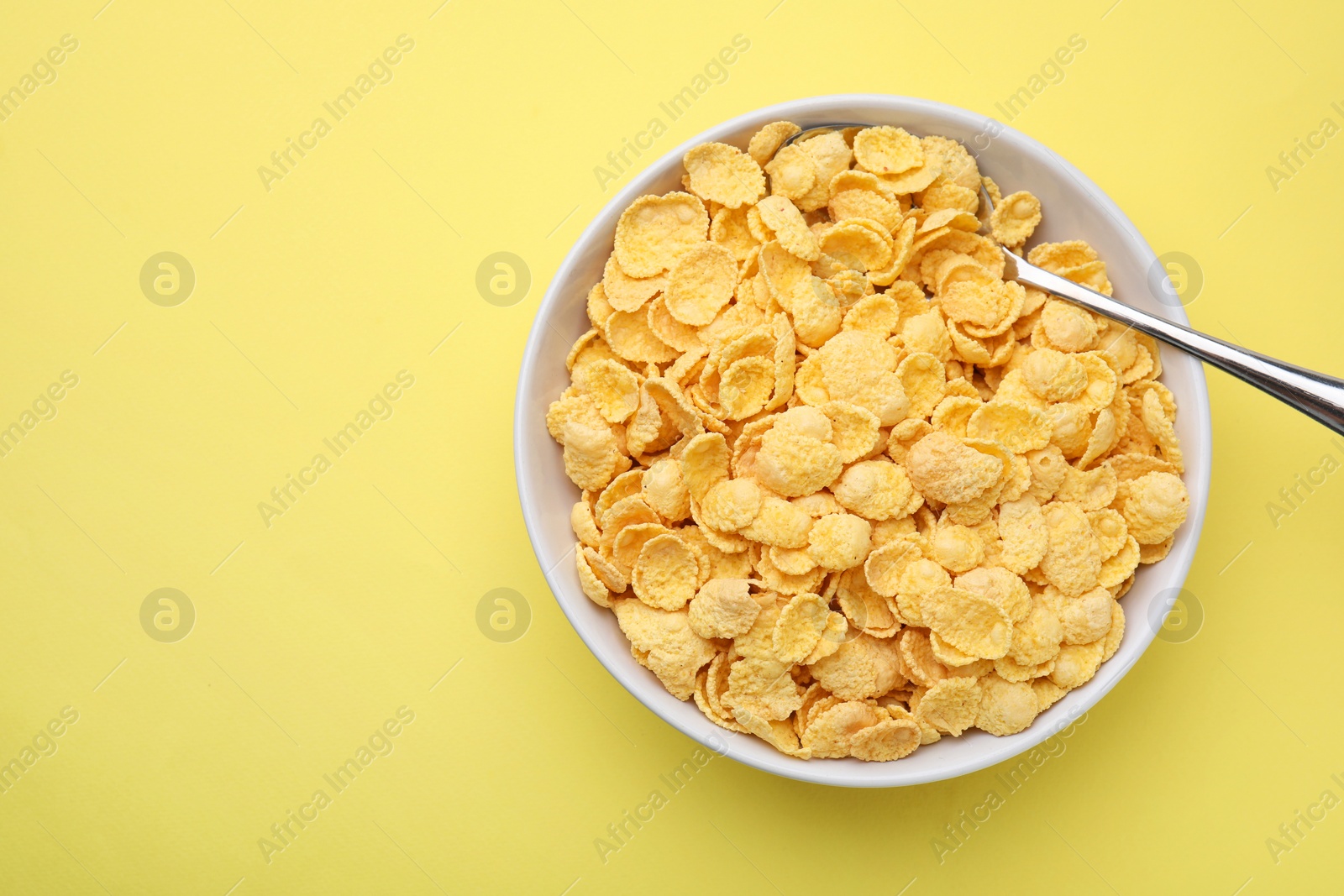 Photo of Breakfast cereal. Tasty corn flakes in bowl and spoon on yellow table, top view. Space for text