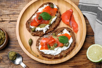 Photo of Delicious sandwiches with cream cheese, salmon, capers and pesto served on wooden table, flat lay