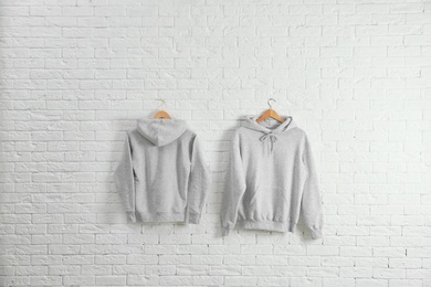 Photo of New hoodie sweaters with hangers on brick wall. Mockup for design