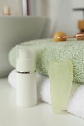 Jade gua sha tool, cosmetic product and folded towels on white countertop, closeup