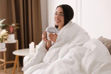 Woman covered in blanket holding cup of drink on sofa