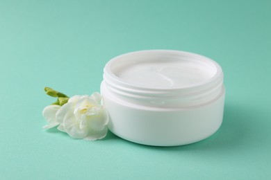 Photo of Moisturizing cream in open jar and freesia flower on turquoise background. Body care product