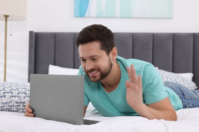 Photo of Happy man greeting someone during video chat via laptop in bedroom