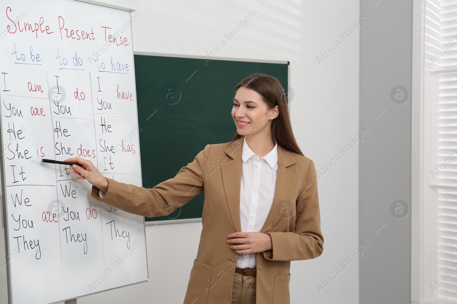Photo of English teacher giving lesson on simple present tense near whiteboard in classroom