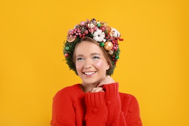 Beautiful young woman wearing Christmas wreath on yellow background