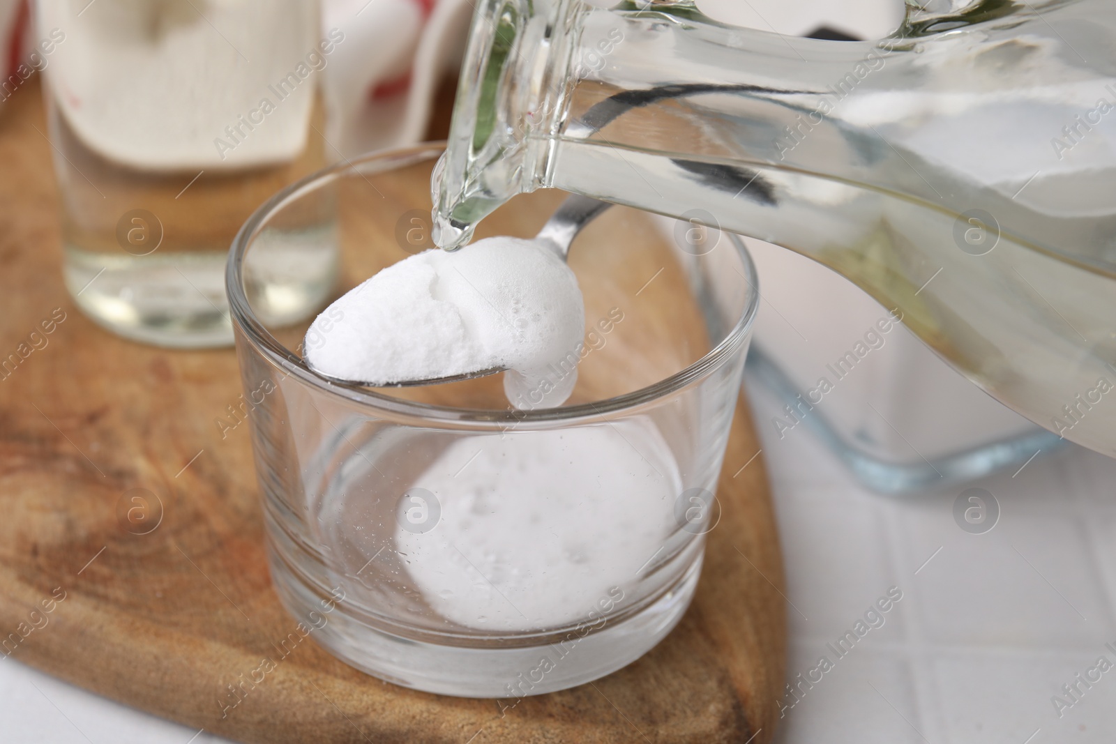 Photo of Pouring vinegar into spoon with baking soda over glass bowl at white table, closeup