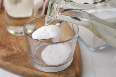 Pouring vinegar into spoon with baking soda over glass bowl at white table, closeup