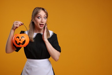 Photo of Emotional woman in scary maid costume with pumpkin bucket on orange background, space for text. Halloween celebration