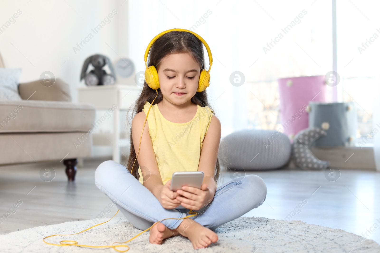 Photo of Cute child with headphones and mobile phone on floor indoors