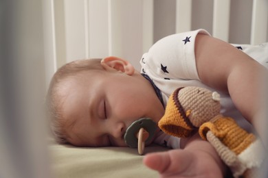 Photo of Adorable little baby with pacifier and toy sleeping in crib, closeup