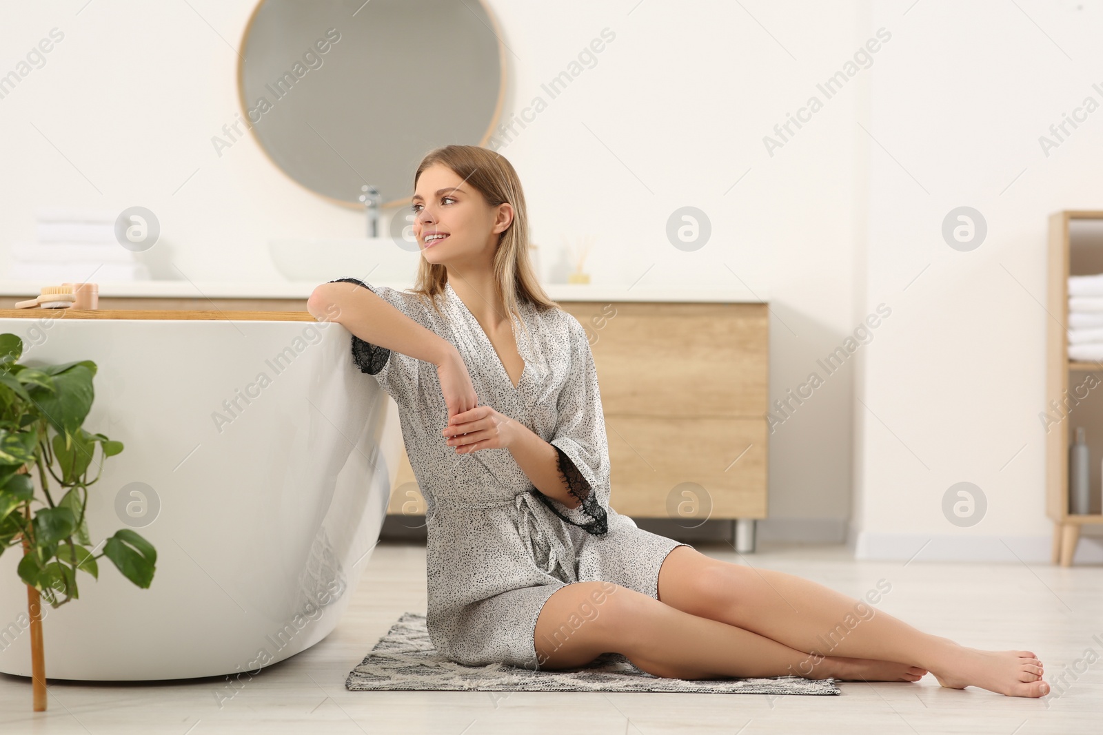 Photo of Beautiful happy woman in stylish robe sitting on floor near bath tub indoors