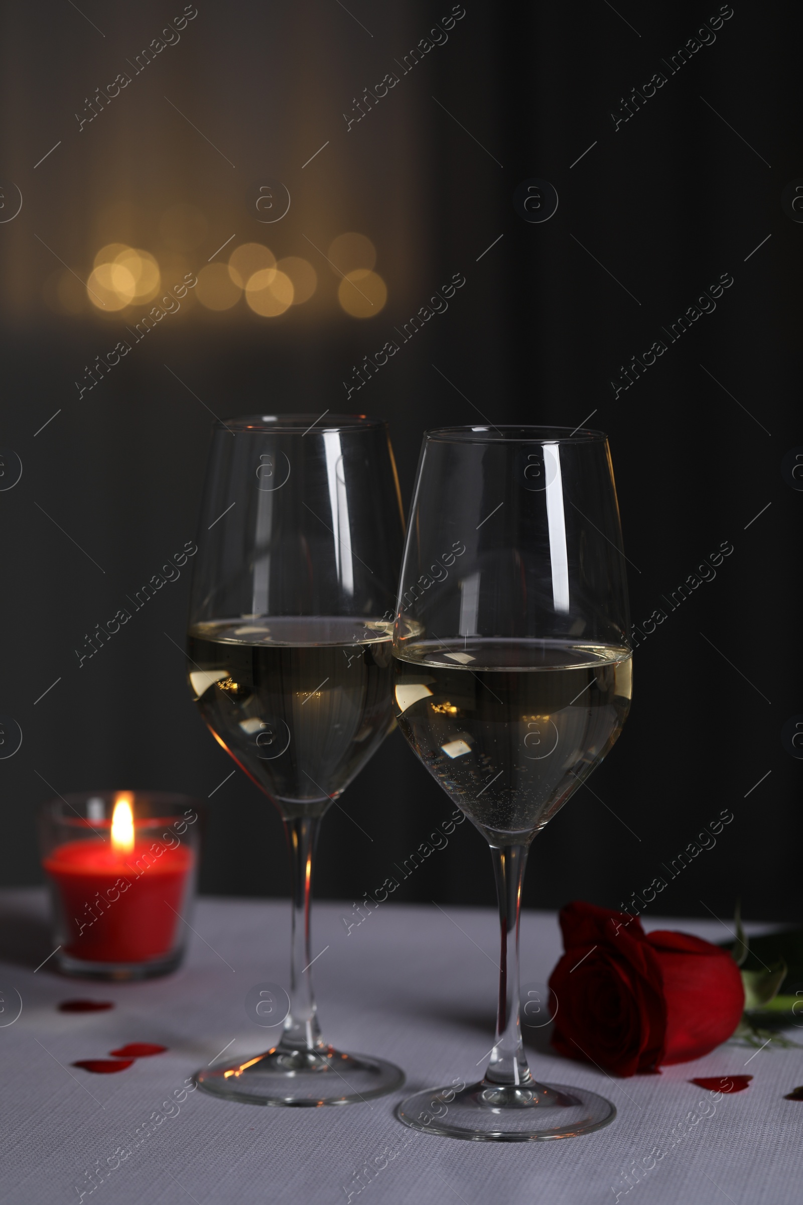 Photo of Glasses of white wine, burning candle and rose flower on grey table against blurred lights. Romantic atmosphere