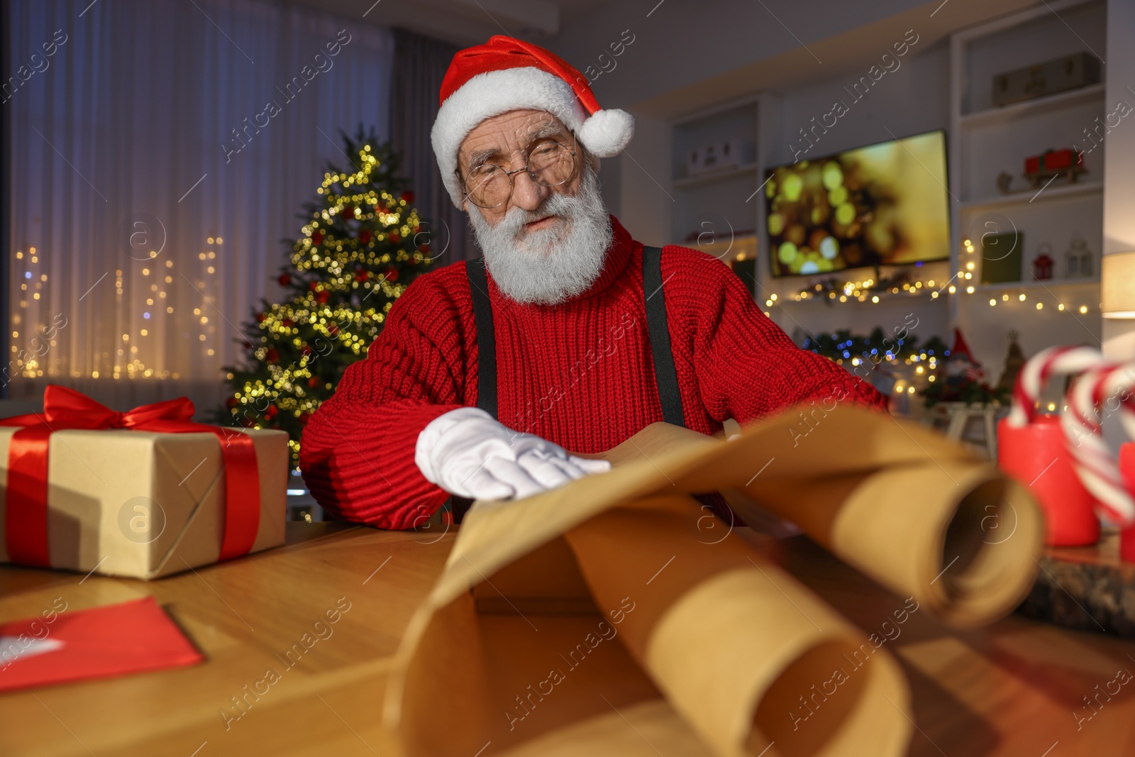 Photo of Santa Claus wrapping gift at his workplace in room decorated for Christmas