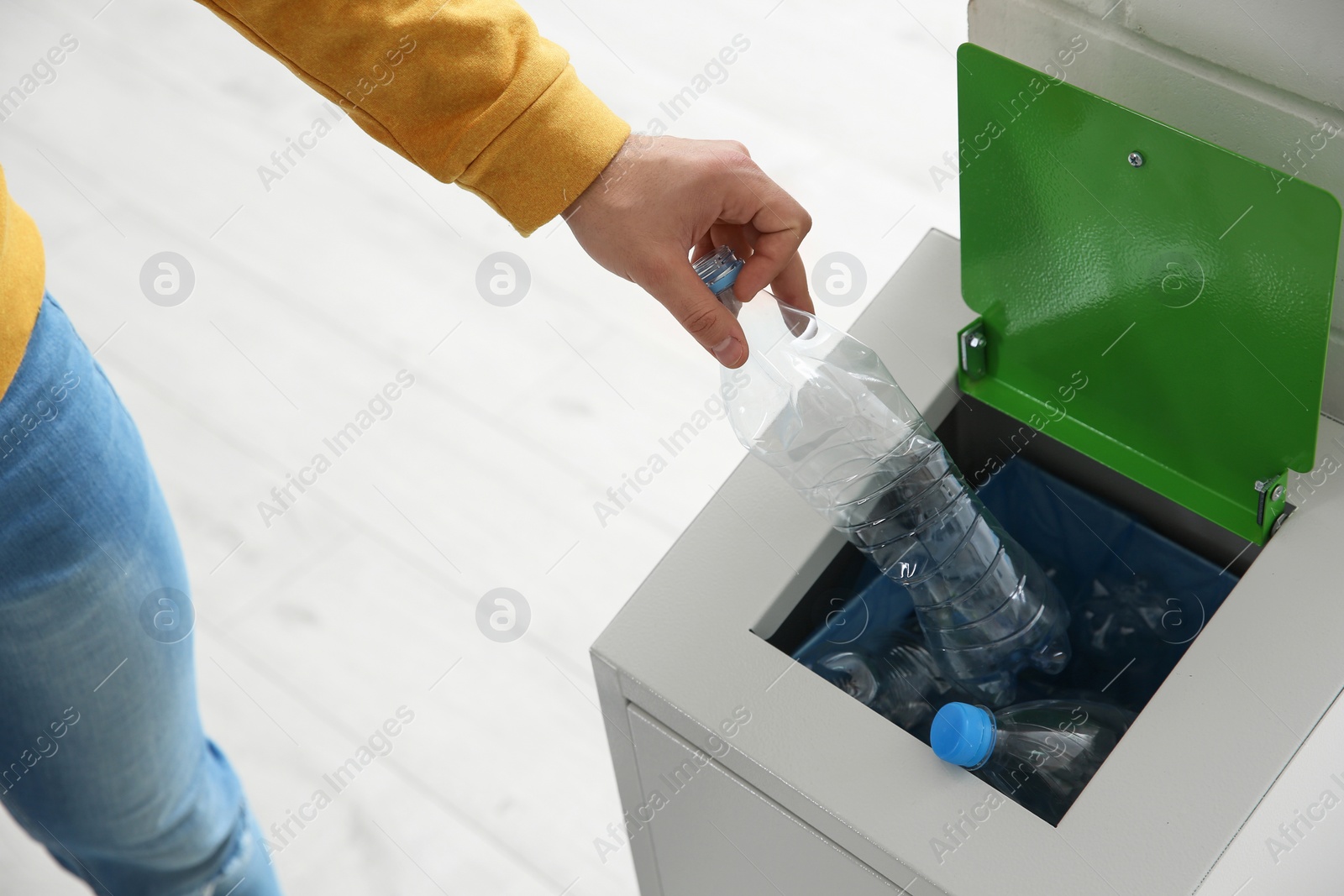 Photo of Young man throwing empty plastic bottle in metal bin indoors, closeup. Waste recycling