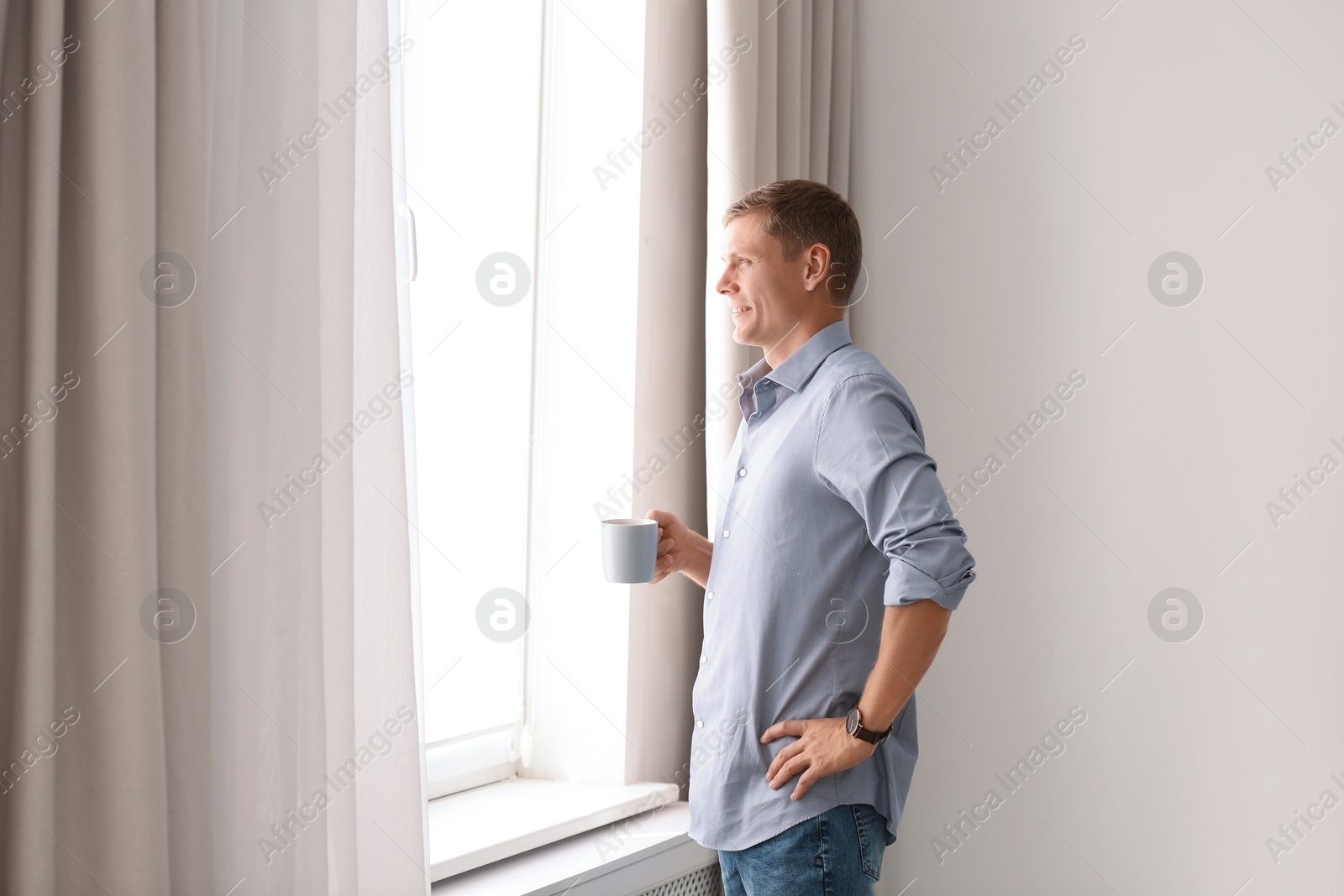 Photo of Mature man with cup of drink near window with open curtains at home. Space for text