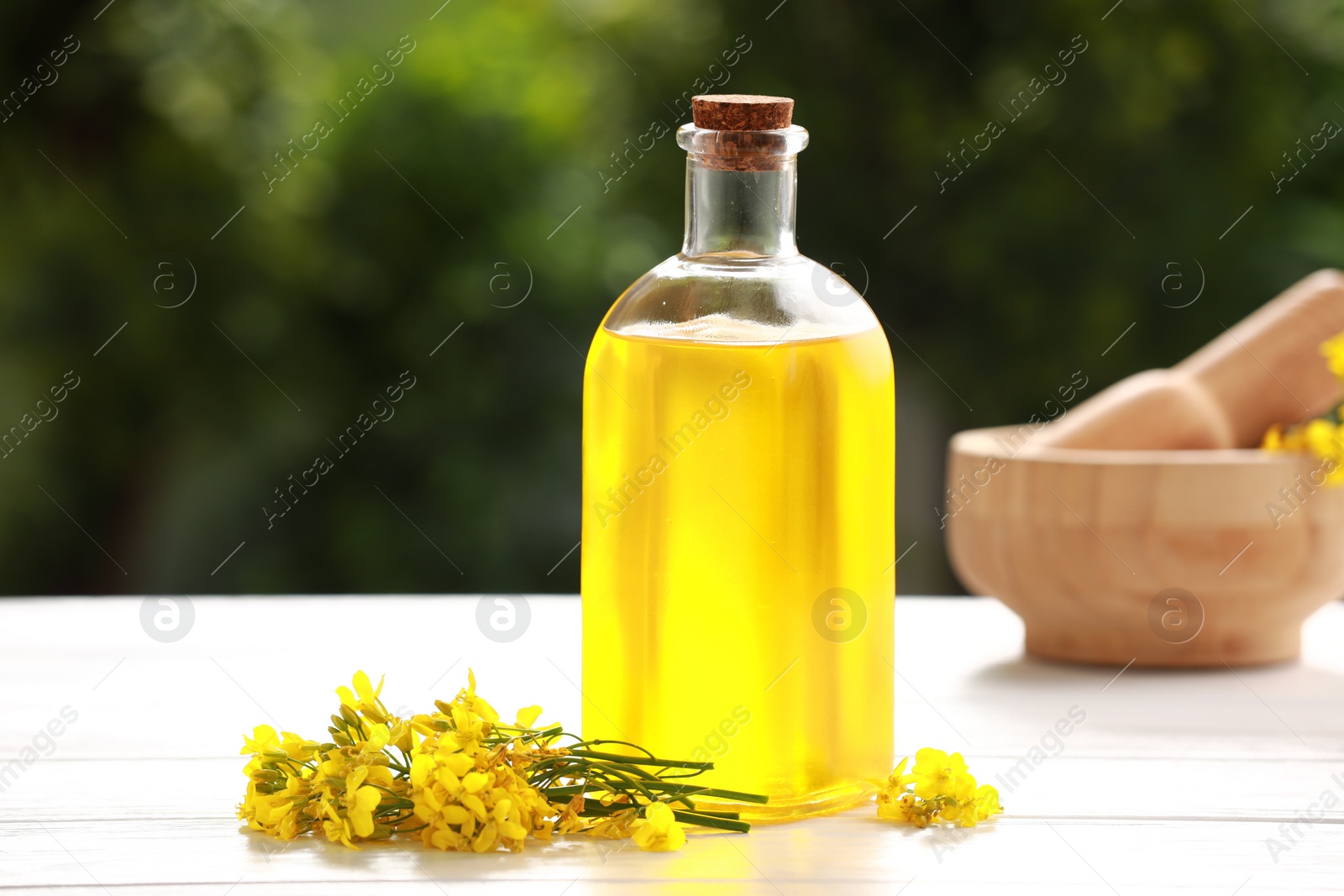 Photo of Rapeseed oil in glass bottle and yellow flowers on white wooden table outdoors