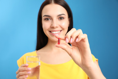 Photo of Young woman with glass of water and vitamin capsule against light blue background, focus on hand