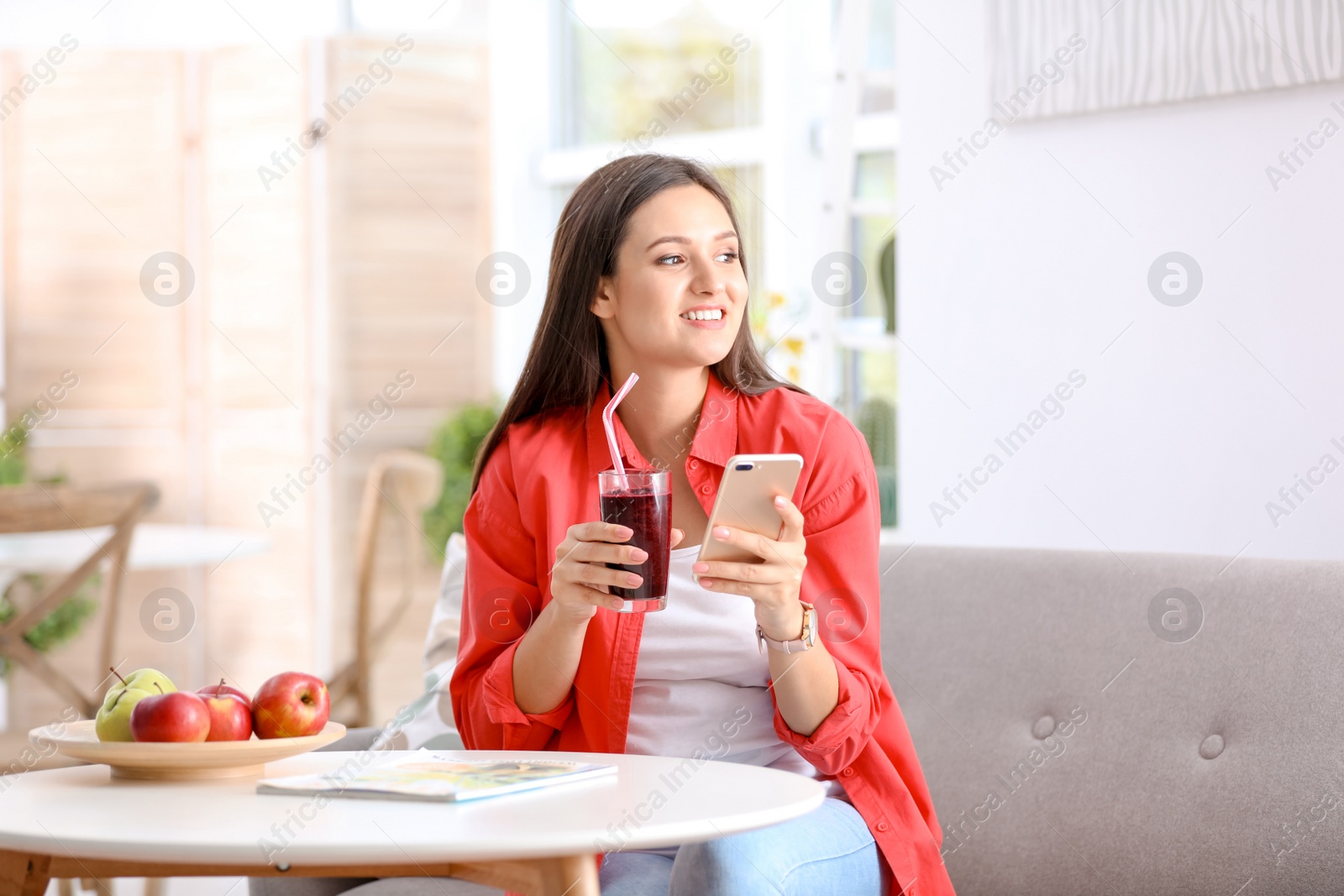 Photo of Young woman using mobile phone while drinking tasty healthy smoothie at table, indoors