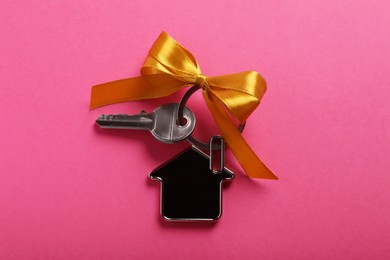 Key with trinket in shape of house and bow on pink background, top view. Housewarming party