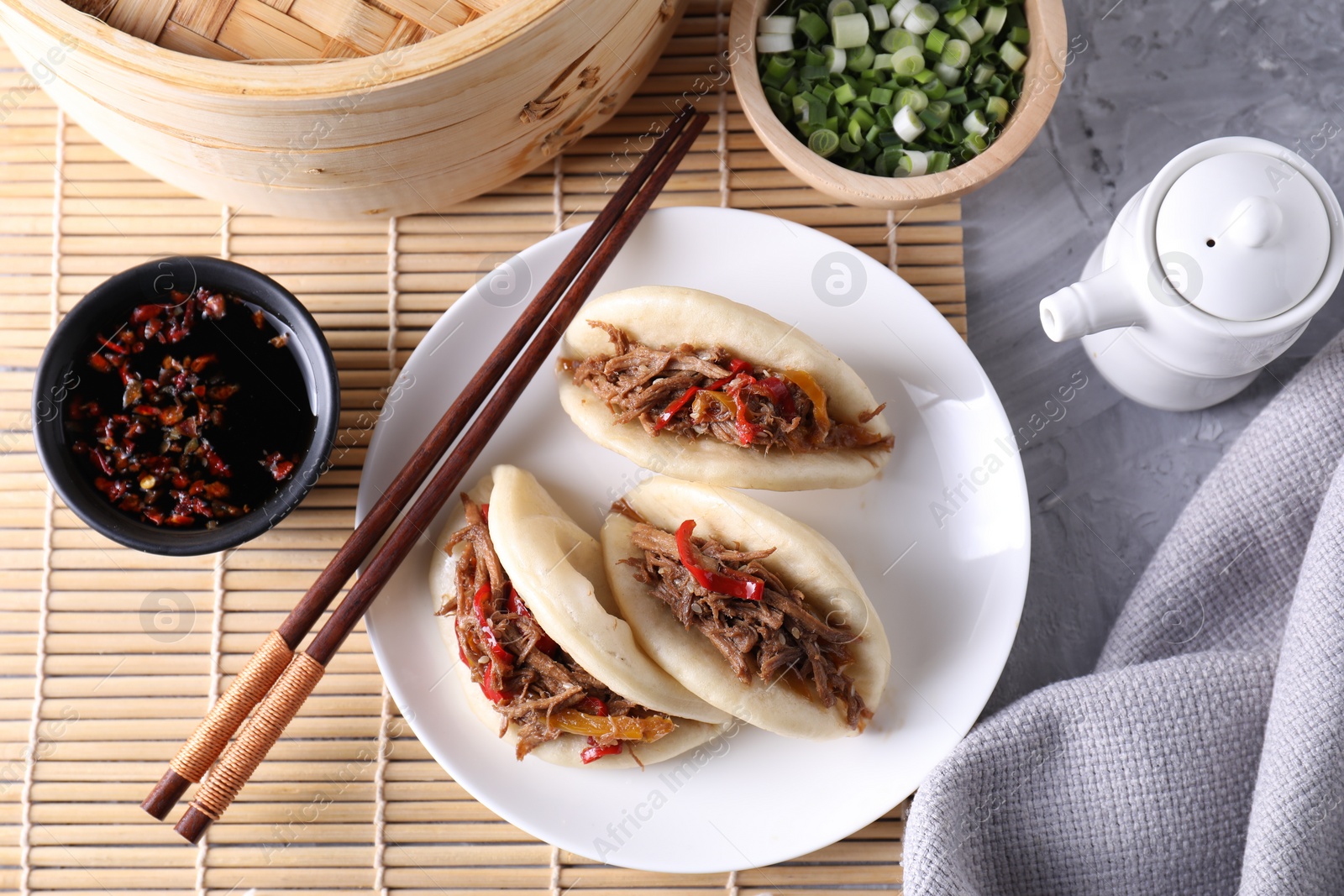 Photo of Delicious gua bao (pork belly buns), chopsticks, green onion and sauces on grey textured table, flat lay