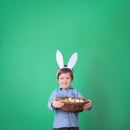 Cute little boy wearing bunny ears with basket full of dyed Easter eggs on green background