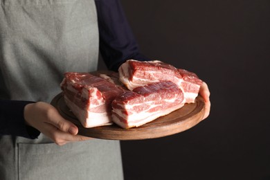 Woman holding wooden board with pieces of raw pork belly on black background, closeup