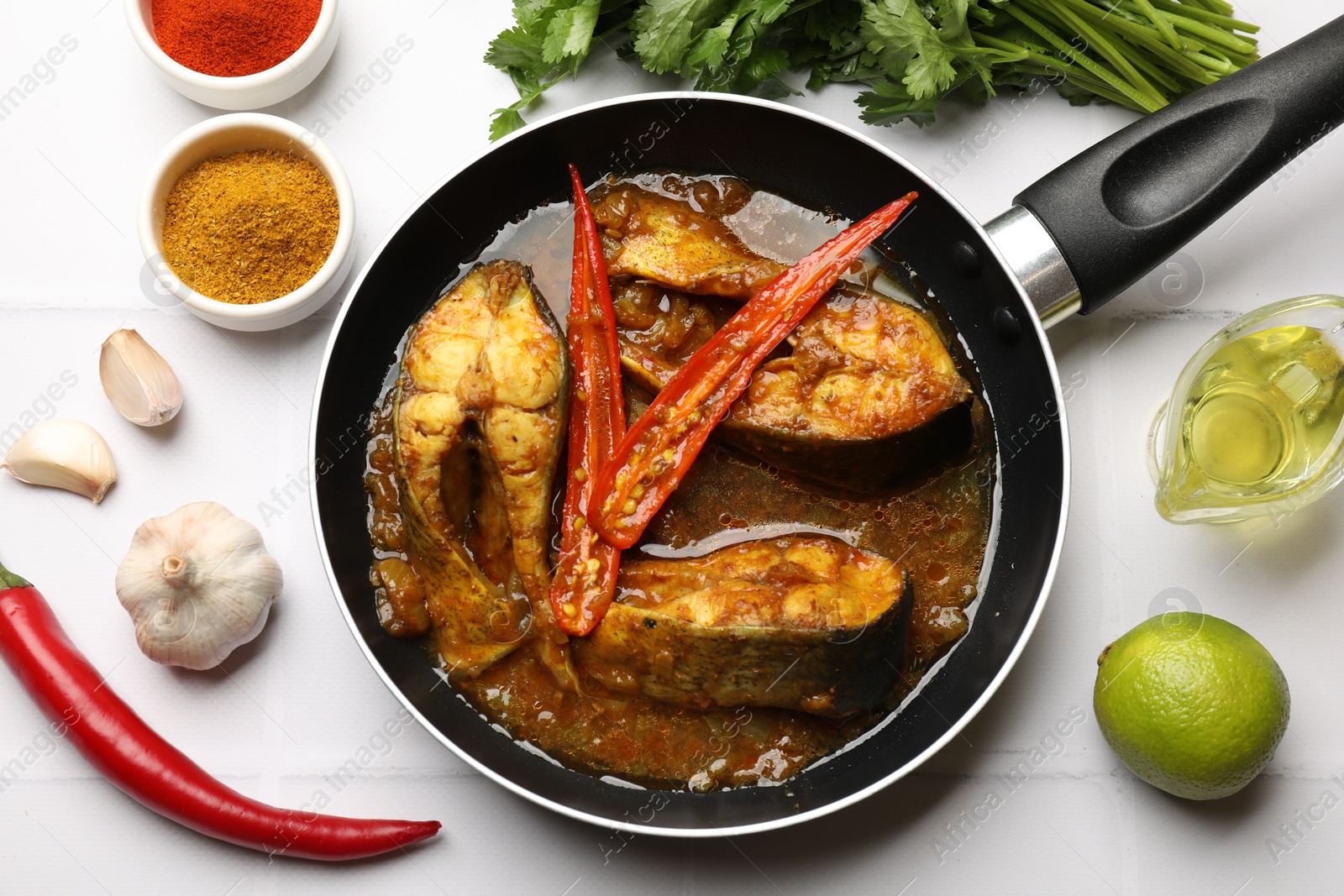 Photo of Tasty fish curry in frying pan and ingredients on white tiled table, flat lay. Indian cuisine