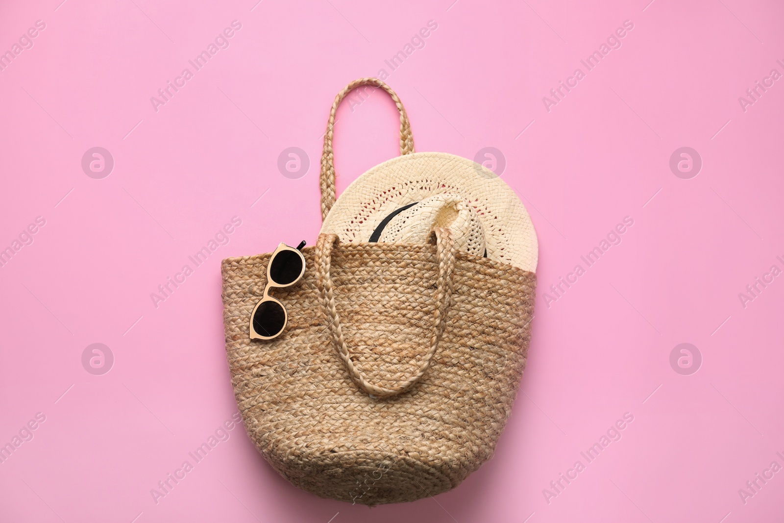 Photo of Elegant woman's straw bag with hat and sunglasses on pink background, top view