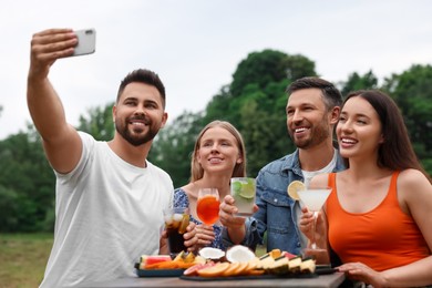 Photo of Happy friends with glasses of cocktails taking selfie at table outdoors