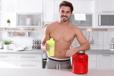 Photo of Young shirtless man holding protein shake in bottle near table with jar of powder at home