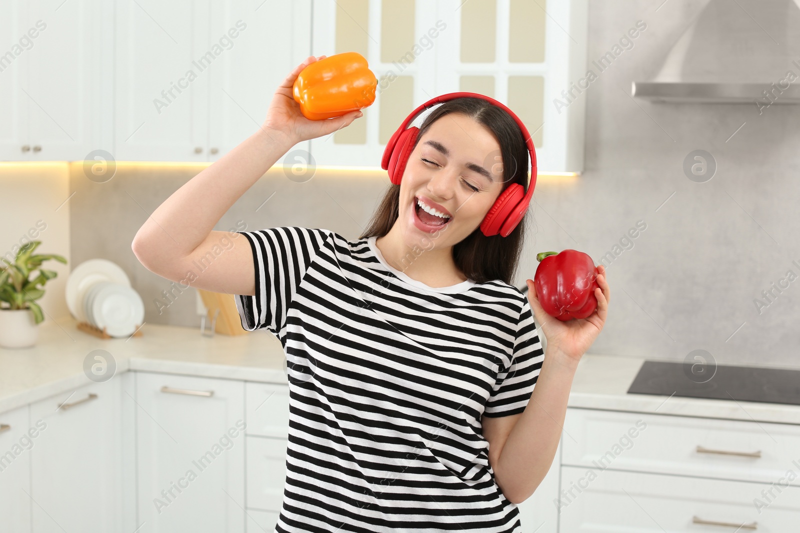 Photo of Happy woman in headphones listening music and dancing with bell peppers in kitchen