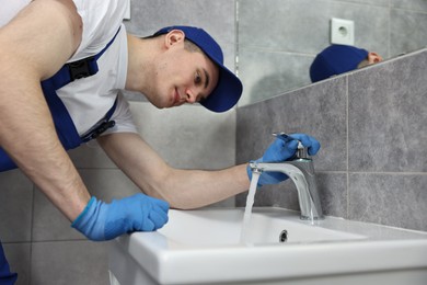 Young plumber wearing gloves examining faucet in bathroom