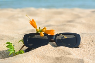 Photo of Stylish sunglasses and tropical flower on sand near sea, closeup