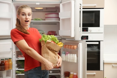 Photo of Young woman with fresh food in paper bag near refrigerator at home