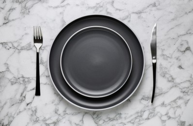 Photo of New dark plates and cutlery on white marble table, flat lay