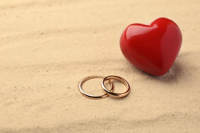 Photo of Honeymoon concept. Two golden rings and red wooden heart on sand