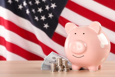 Photo of Piggy bank and money on light wooden table against American flag, space for text