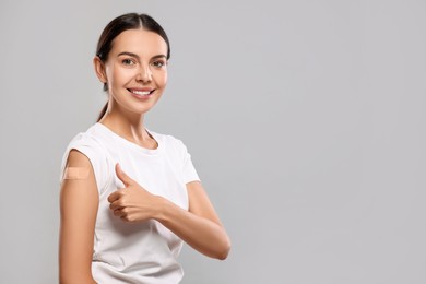 Photo of Woman with sticking plaster on arm after vaccination showing thumbs up against light grey background, space for text
