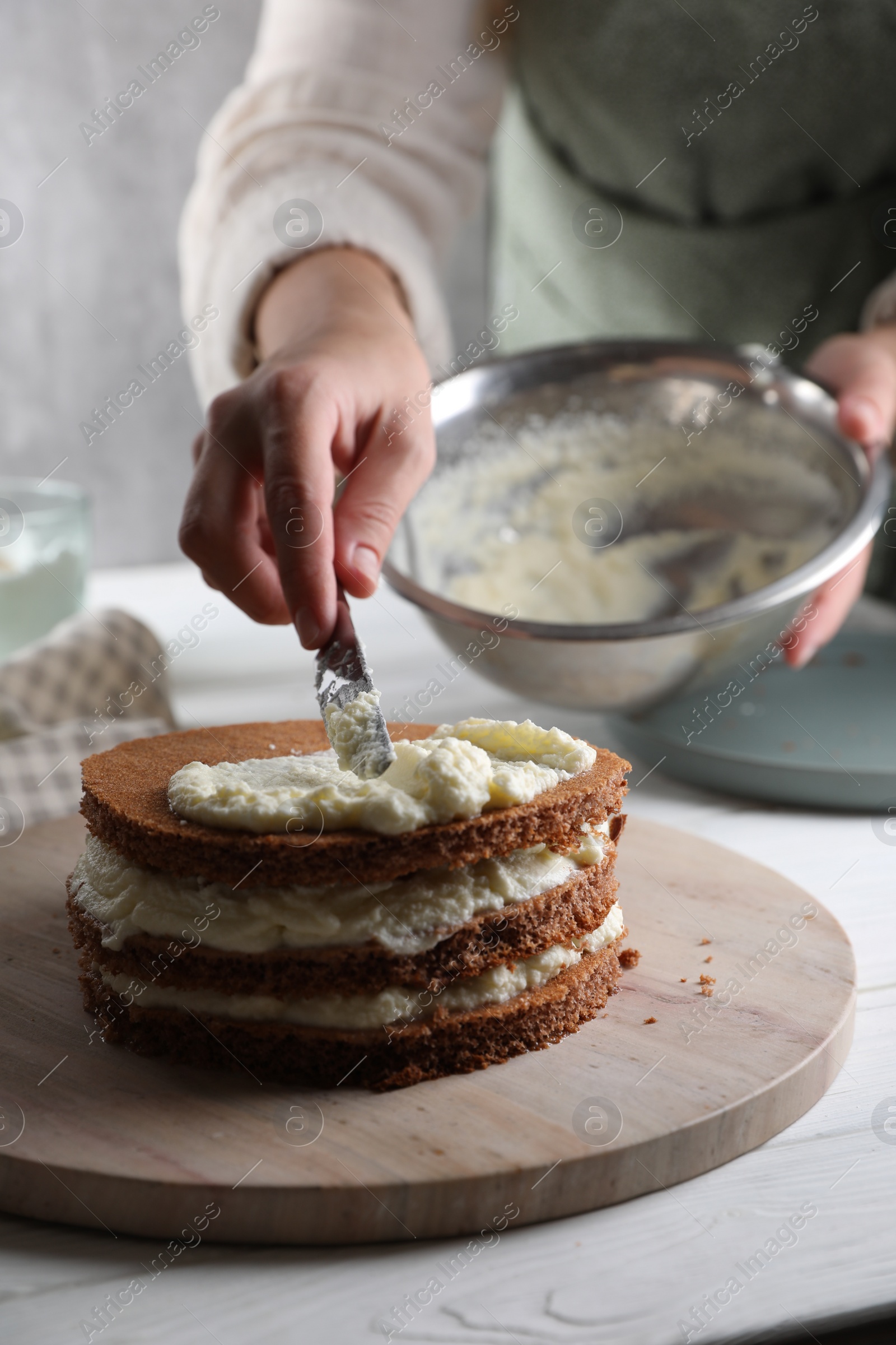 Photo of Woman smearing sponge cake with cream at white wooden table, closeup