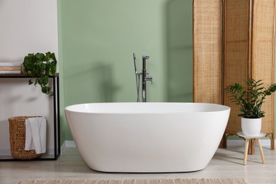 Photo of Modern ceramic tub and beautiful plants near light green wall in bathroom. Interior design