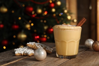 Tasty eggnog with cinnamon, cookies and Christmas baubles on wooden table against blurred festive lights. Space for text