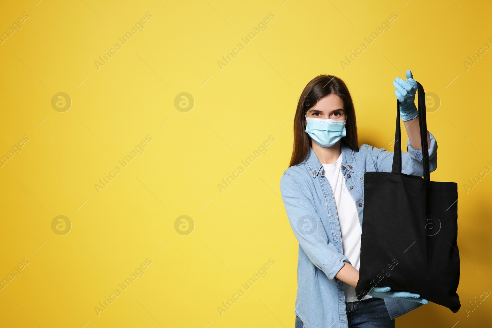 Photo of Female volunteer in protective mask and gloves with black bag on yellow background, space for text. Aid during coronavirus quarantine