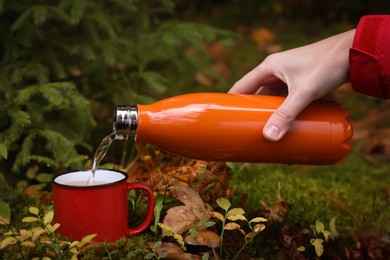 Photo of Woman pouring hot water from thermo bottle into mug on green grass outdoors, closeup