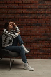 Sad young woman sitting on chair near brick wall indoors