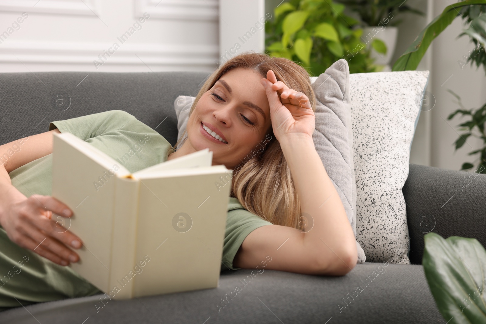 Photo of Woman reading book on sofa in room with houseplants