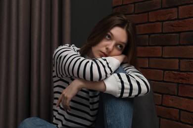 Photo of Sad young woman sitting on chair near brick wall indoors