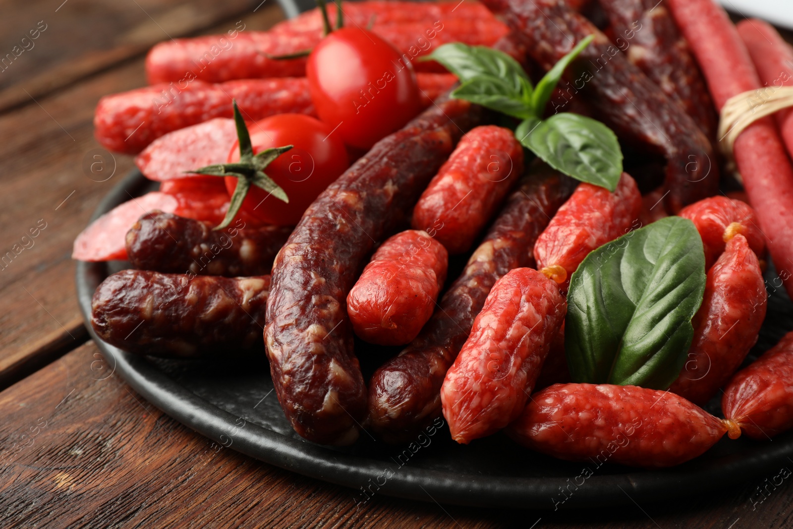 Photo of Different thin dry smoked sausages, basil and tomatoes on wooden table, closeup